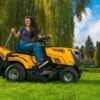 Pani w ogrodzie siedząca na traktorze riwall rlt opróżniająca kosz z trawy w tle staw, dom wierzba i drzewa