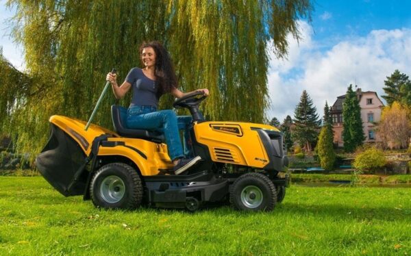 Pani w ogrodzie siedząca na traktorze riwall rlt opróżniająca kosz z trawy w tle staw, dom wierzba i drzewa